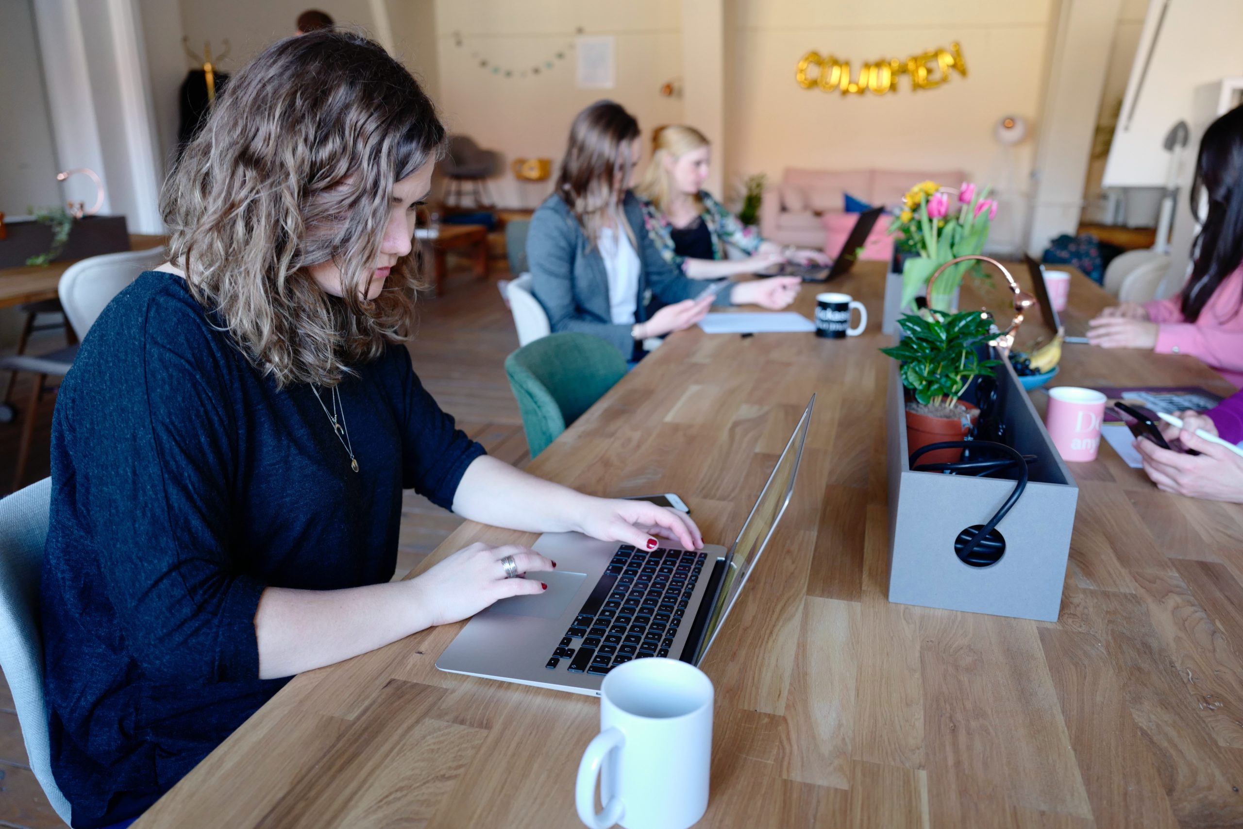 5 women working on laptops around a large desk - decorative image - coworking trends blog
